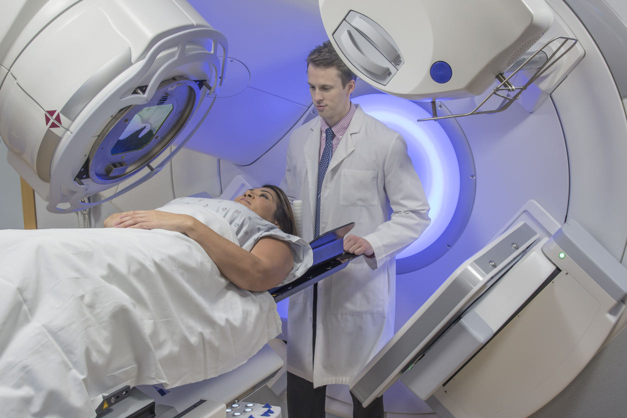 A lady is lying under a radiotherapy machine. A doctor is standing behind her head